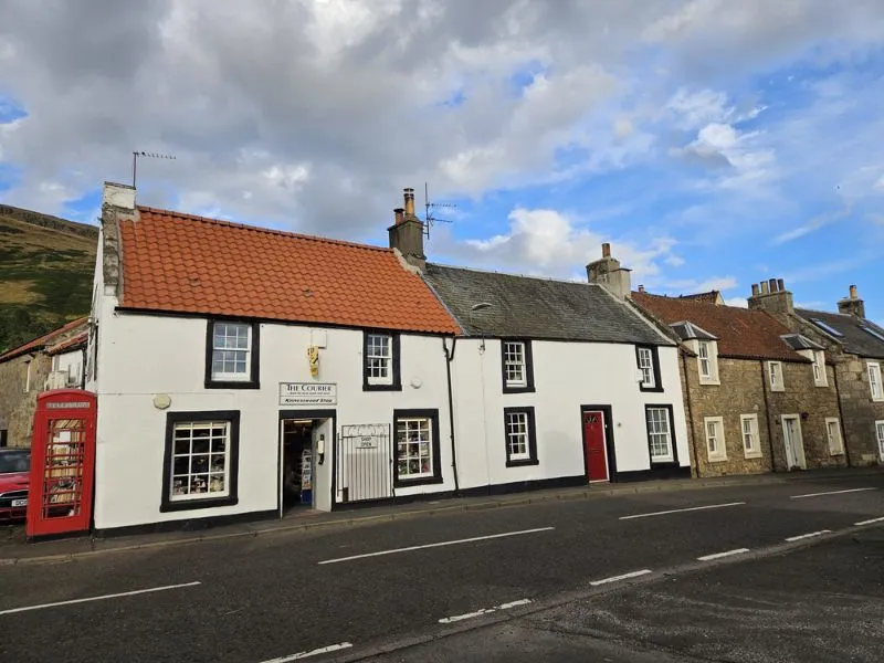 A photograph of Kinnesswod Shop on the main raod that circumnavigates Loch Leven. The shop has a white frontage with black outlines around the windows and a sign above the door saying The Courier and Kinnesswood Shop. There is a red telephone box in the left of the image.